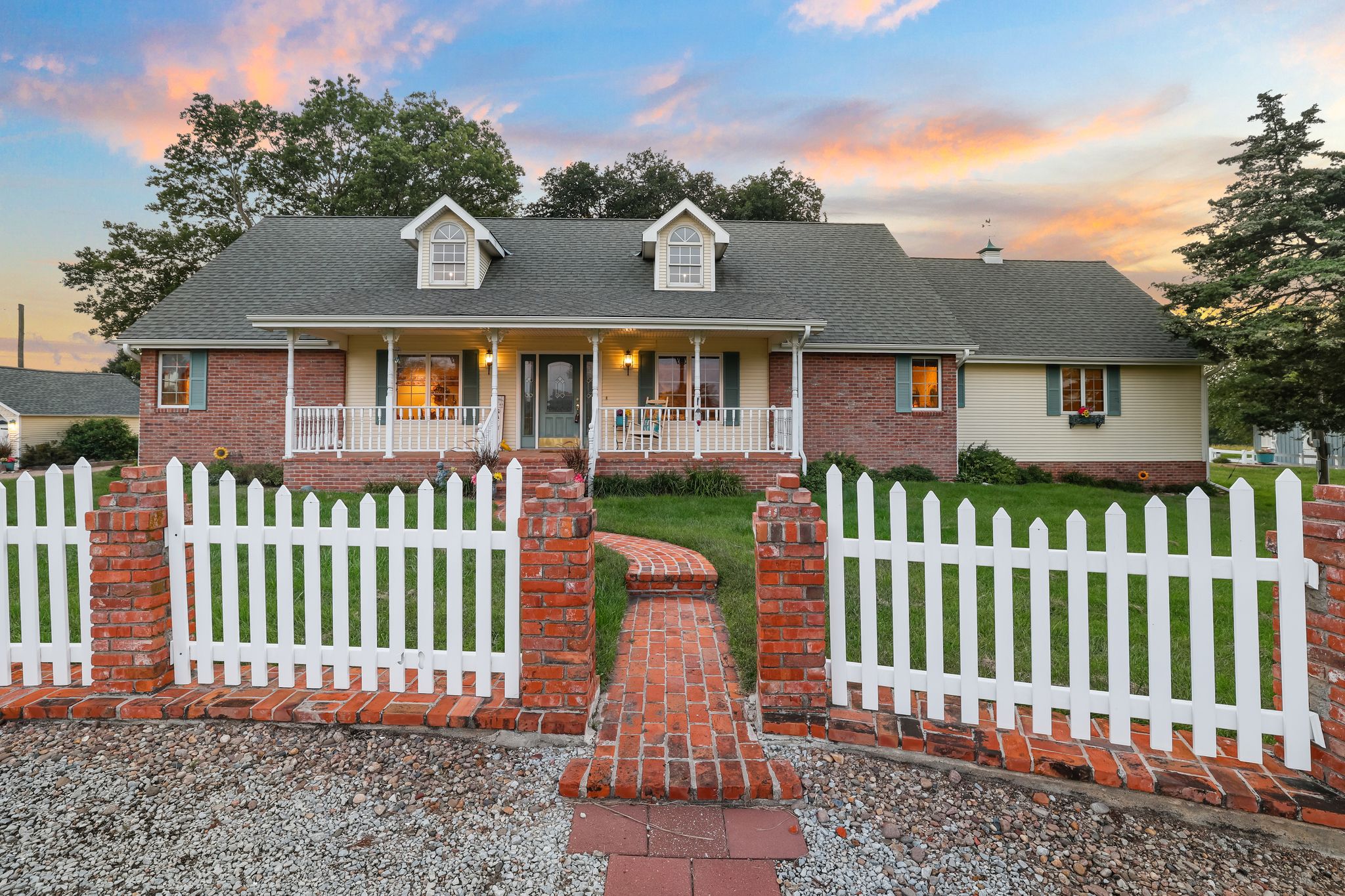 Brick and siding ranch home at dusk with glowing windows, white picket fence, and a sunset sky