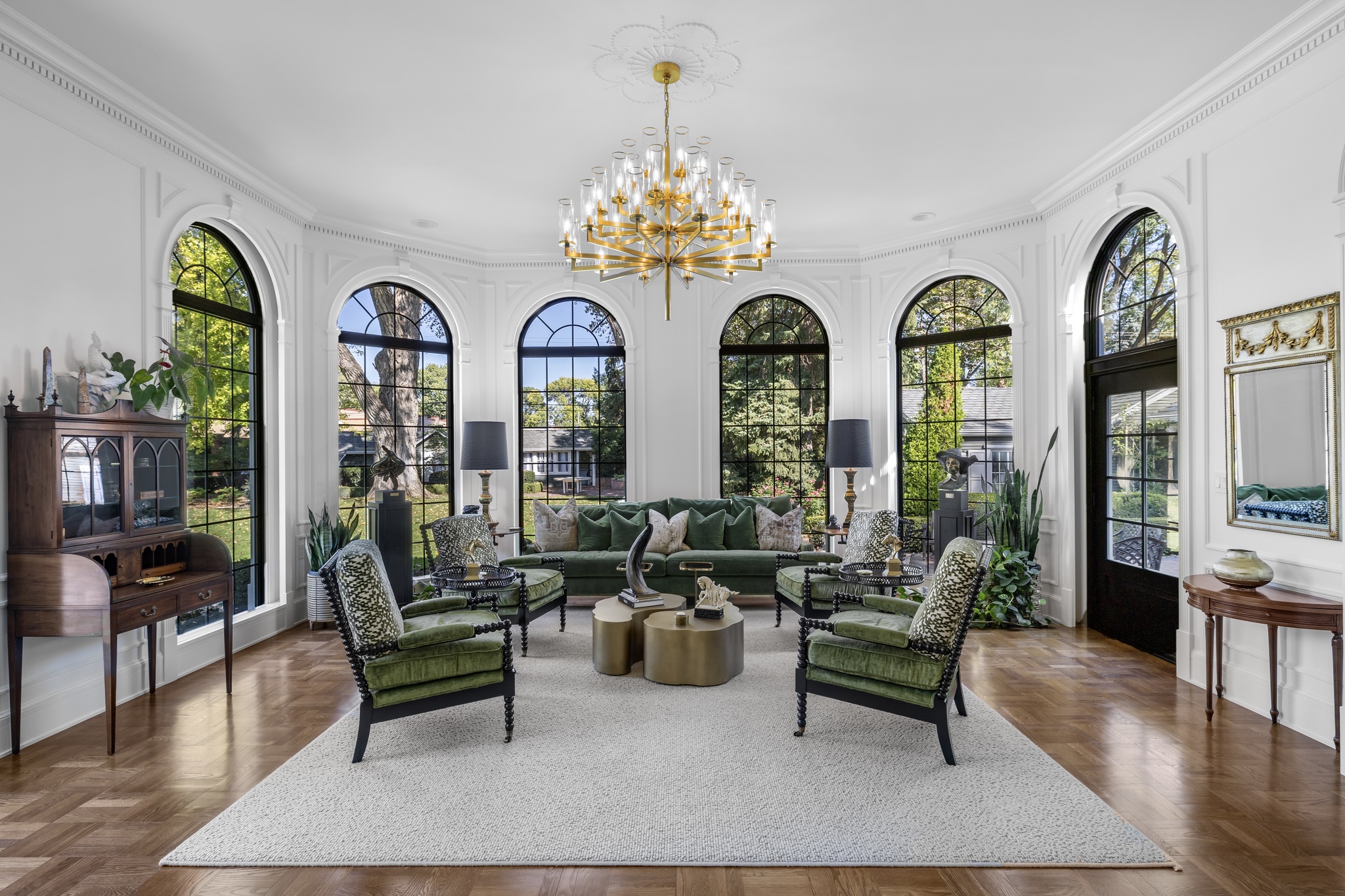 Bright sunroom with arched black-framed windows, a green velvet sofa, and a gold chandelier