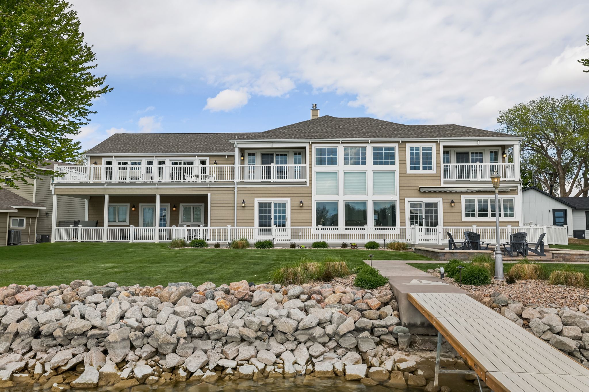 Two-story waterfront home with white decks overlooking a green lawn and rocky shoreline