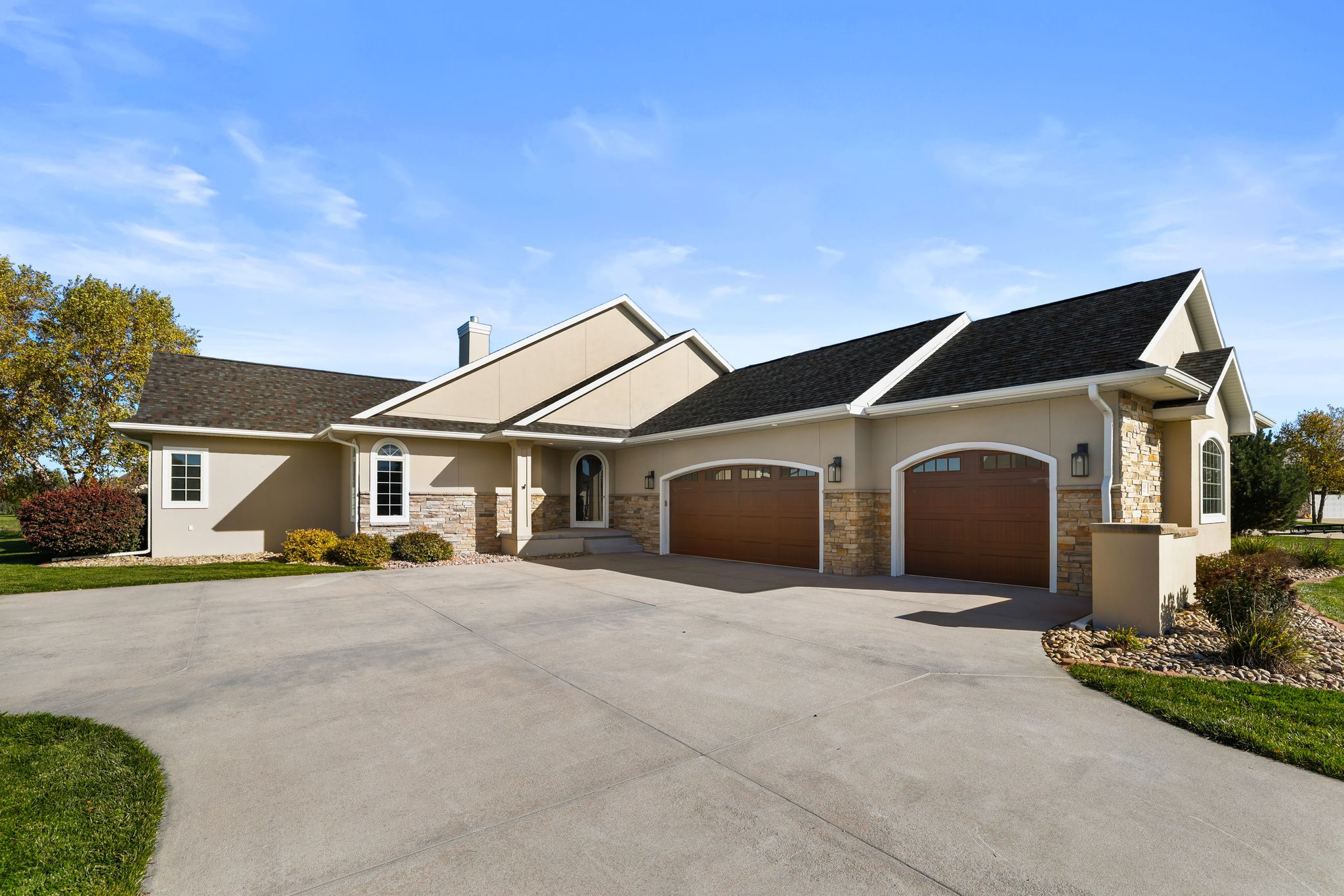 Modern beige home with stone accents, wood garage doors, and a wide concrete driveway under a clear sky