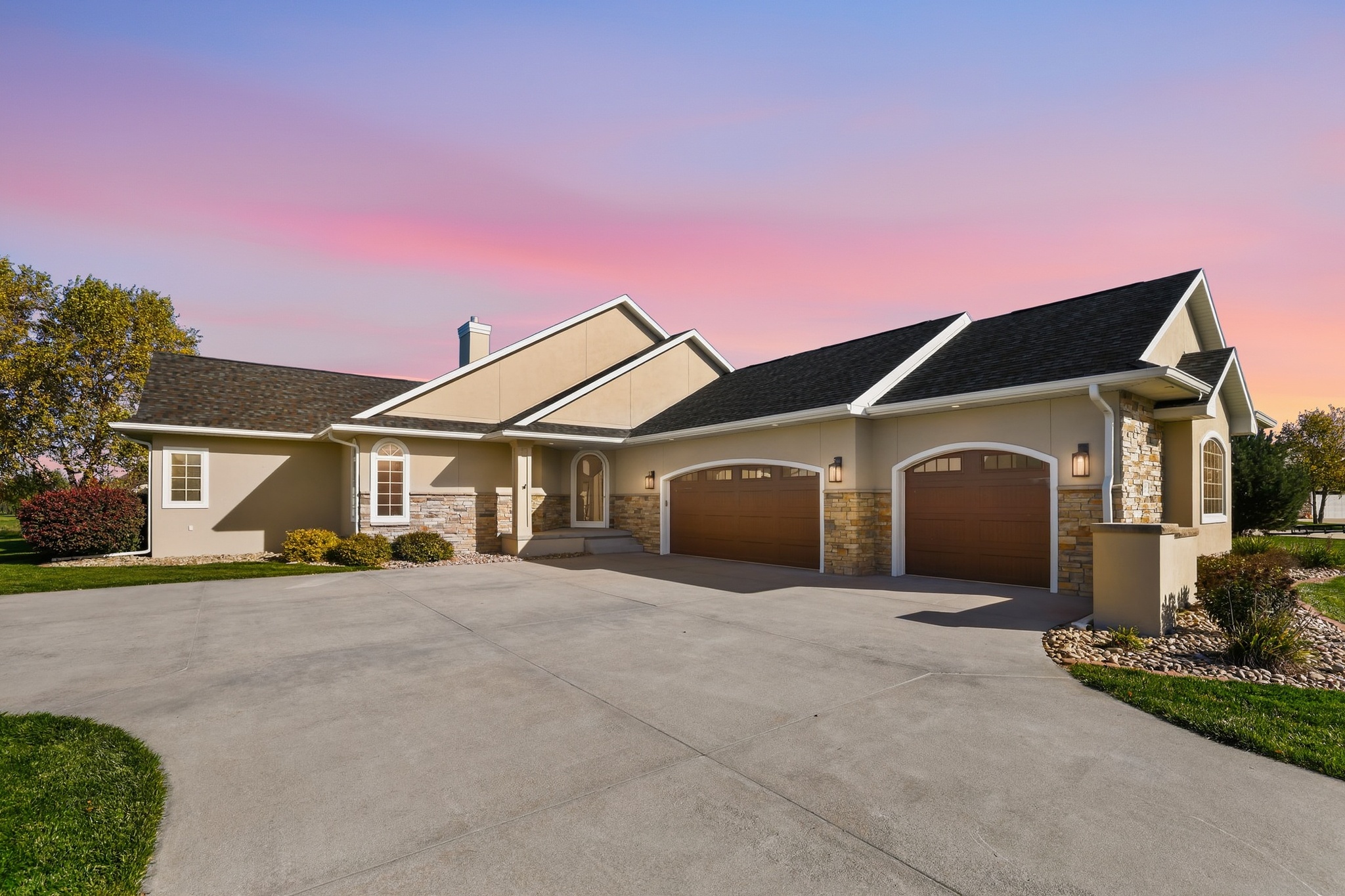 Beige ranch home with stone accents and wood garage doors under a pink and purple sunset sky